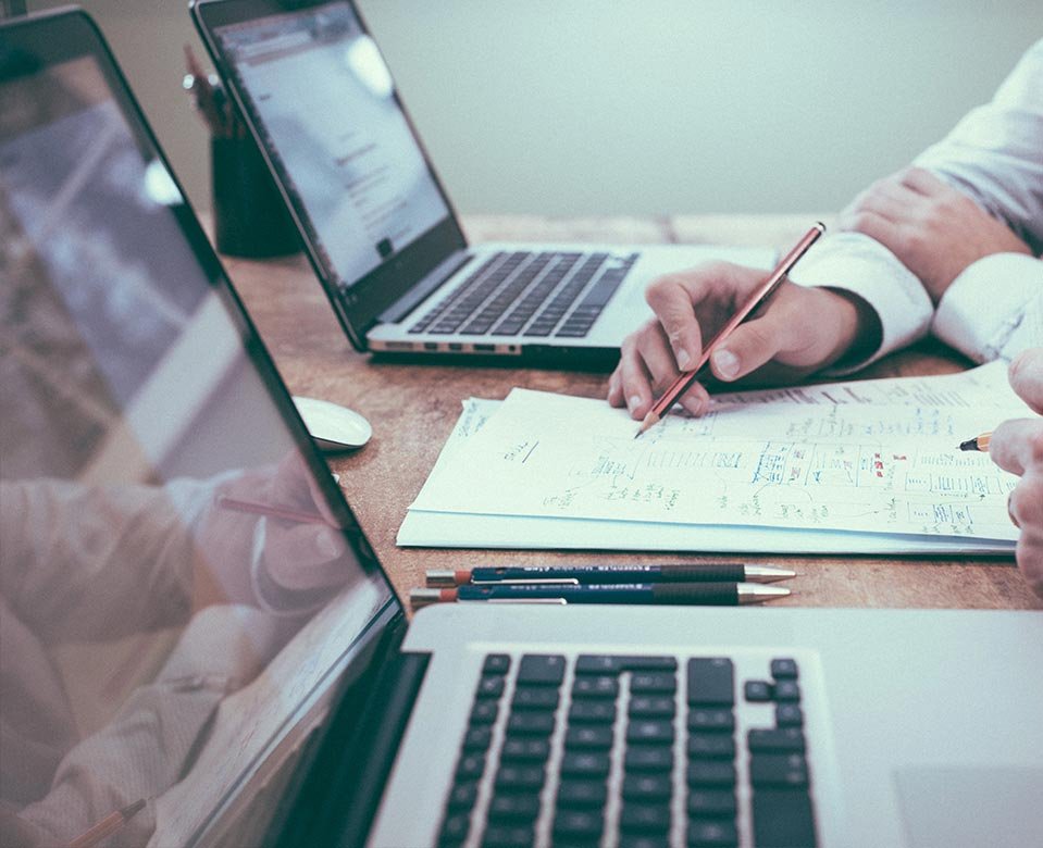 Laptops sobre mesa de trabajo con apuntes en una libreta y una mano escribiendo con pluma